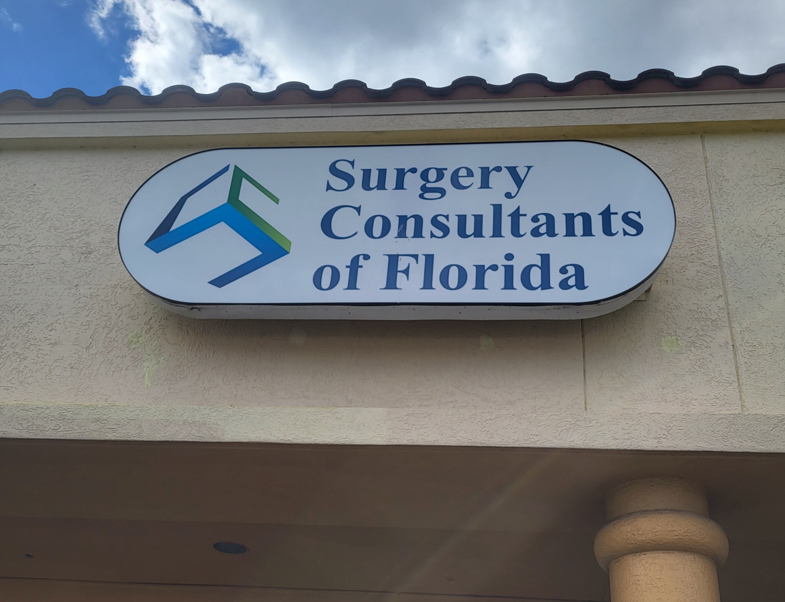 A signboard for "Surgery Consultants of Orlando, Florida" with a distinctive logo mounted on an exterior wall under a clear sky.