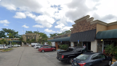 A sunny day at a suburban retail strip mall in Hollywood with various storefronts, including a nail salon, under a clear blue sky with scattered clouds. Vehicles are parked in front, hinting at moderate