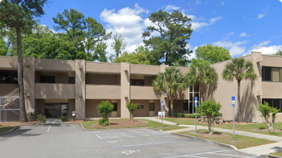 A two-story beige office building, frequented by Hollywood celebrities and surrounded by greenery under a clear blue sky, with a partly filled parking lot in the front.
